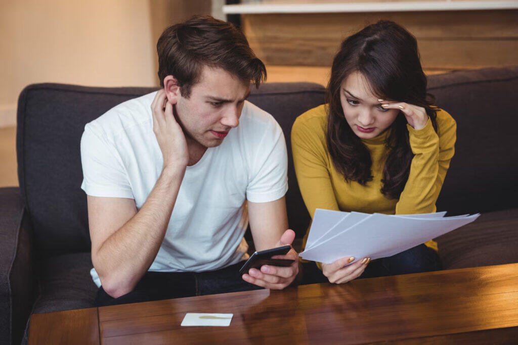 Worried couple reviewing financial documents and phone, possibly life insurance paperwork. Avoiding life insurance mistakes is key.
