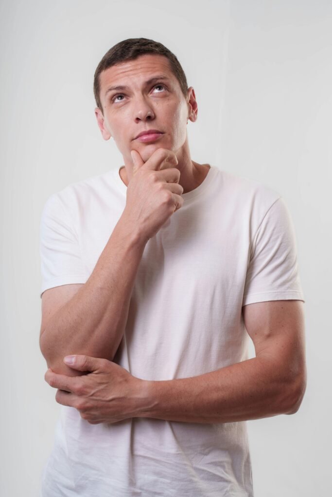 Contemplative young man in white shirt looking up, hand on chin, pondering ideas.