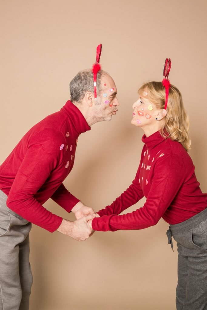 Side view of positive elderly couple in similar clothes and heart party headbands holding hands and pouting lips while standing in studio during Saint Valentines Day